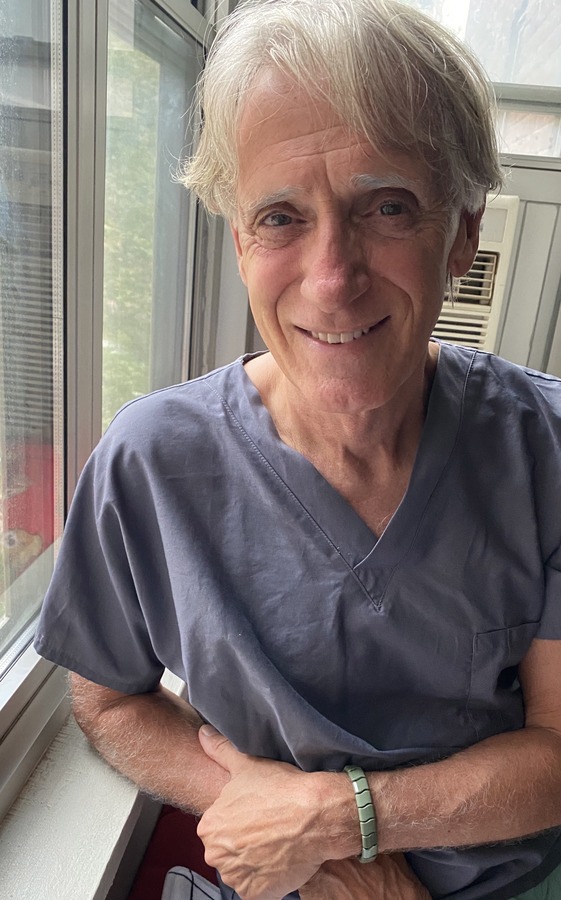 Robert Older man with gray hair wearing gray scrubs sits by a window, smiling at the camera.