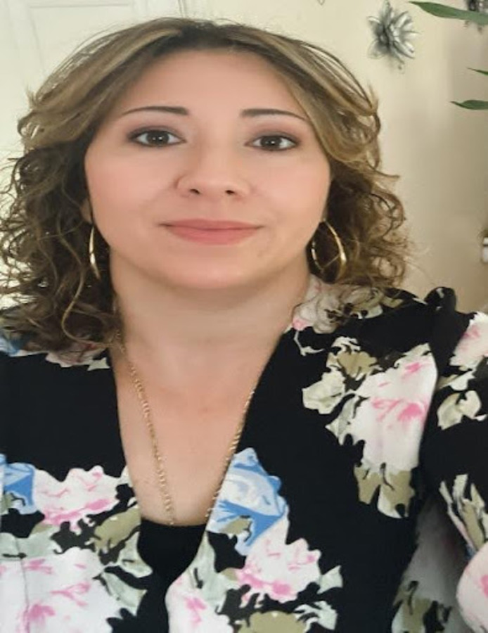 Natalia Medero A woman with shoulder-length curly hair, wearing hoop earrings and a floral-patterned top, looks at the camera.