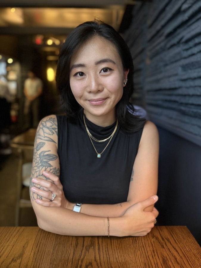 Samantha Lui PT, DPT A woman with dark hair, wearing a sleeveless black top and jewelry, sits at a wooden table with her arms crossed, looking at the camera in a dimly lit restaurant.