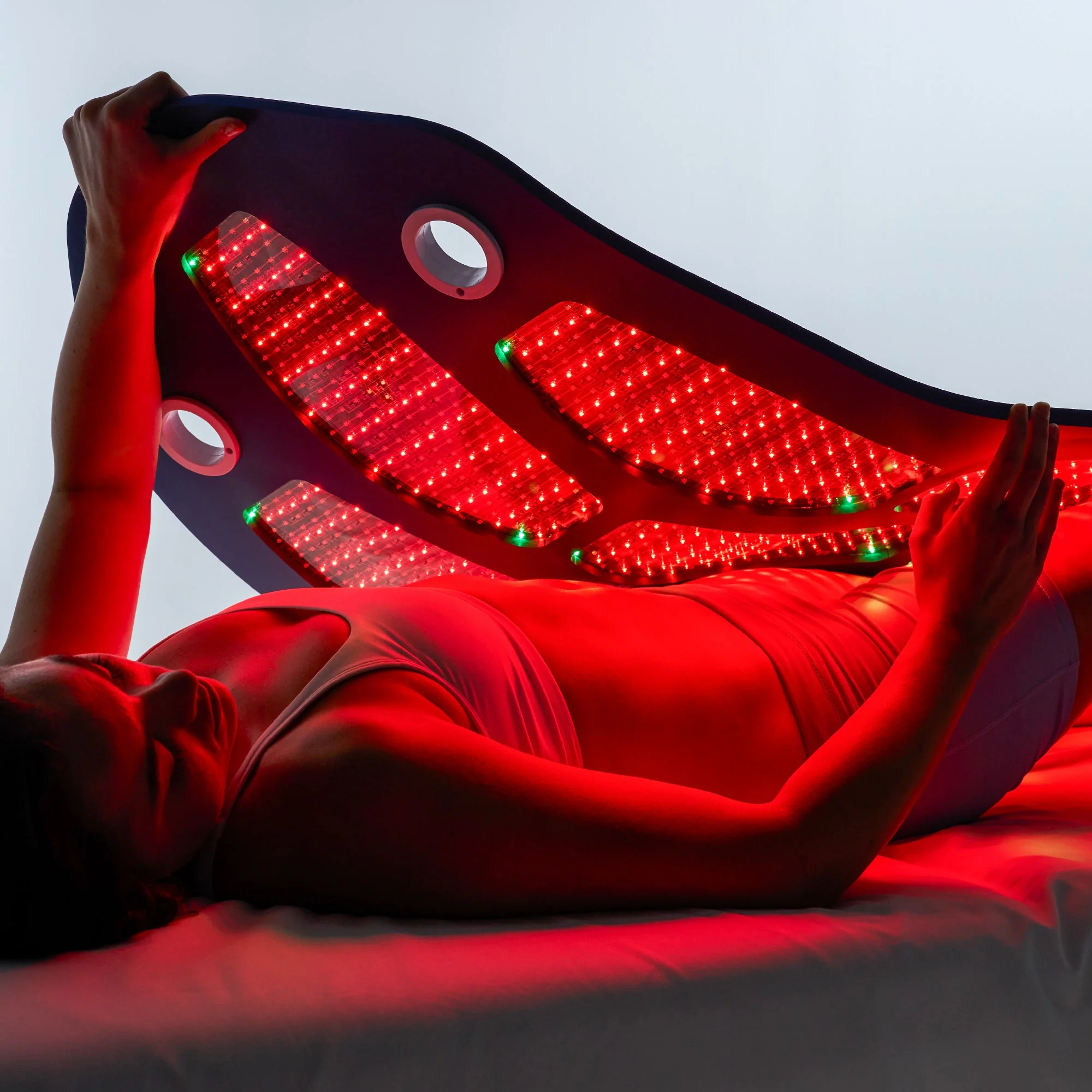 A person lying on a table uses a large red light therapy mat, which is illuminated and covering their torso.
