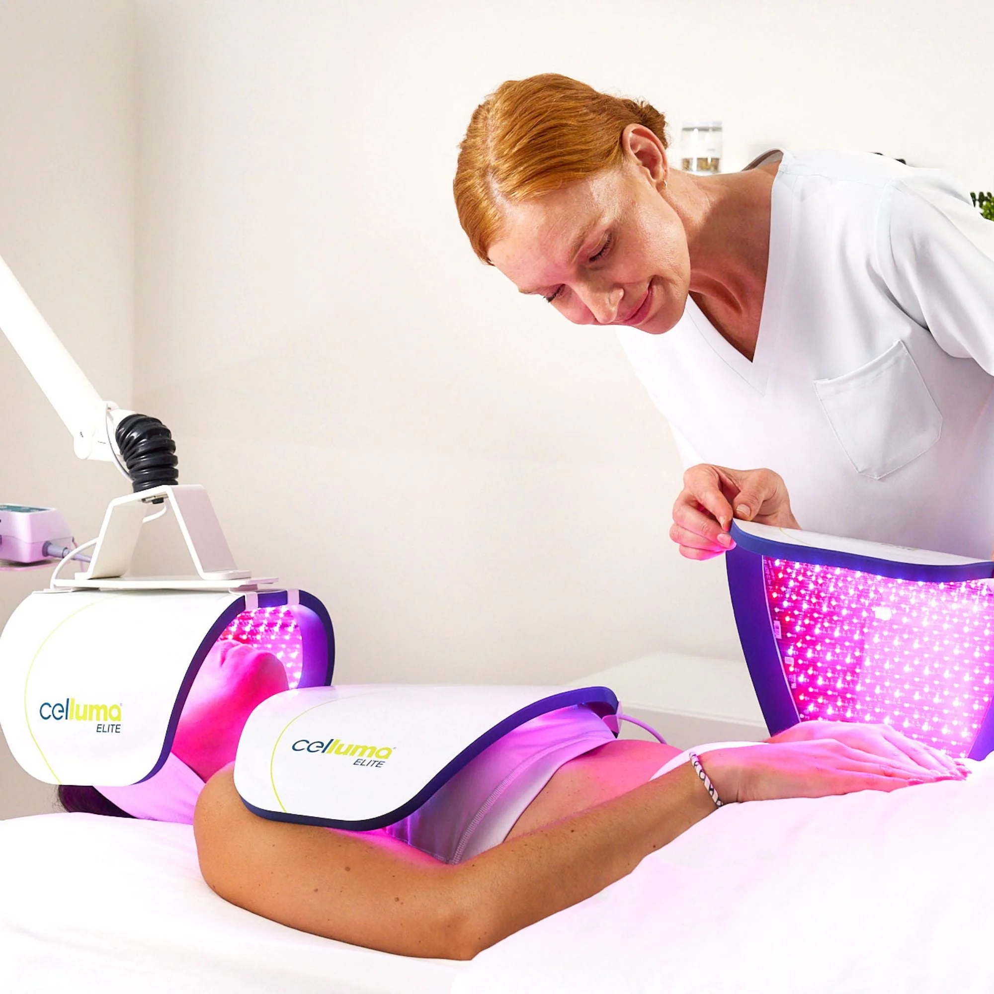 A person receives a light therapy treatment with LED panels on their body while a practitioner adjusts the equipment in a clinical setting.