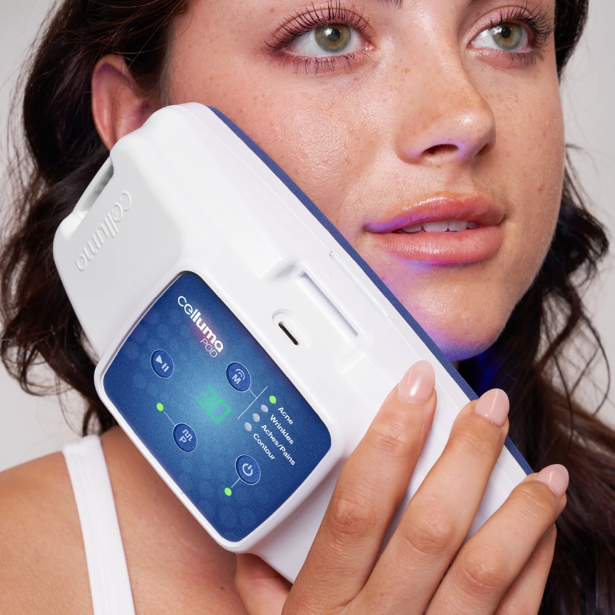 A woman holds a Celluma device against her cheek, using it for light therapy treatment; the device's display shows various mode indicators and a timer.