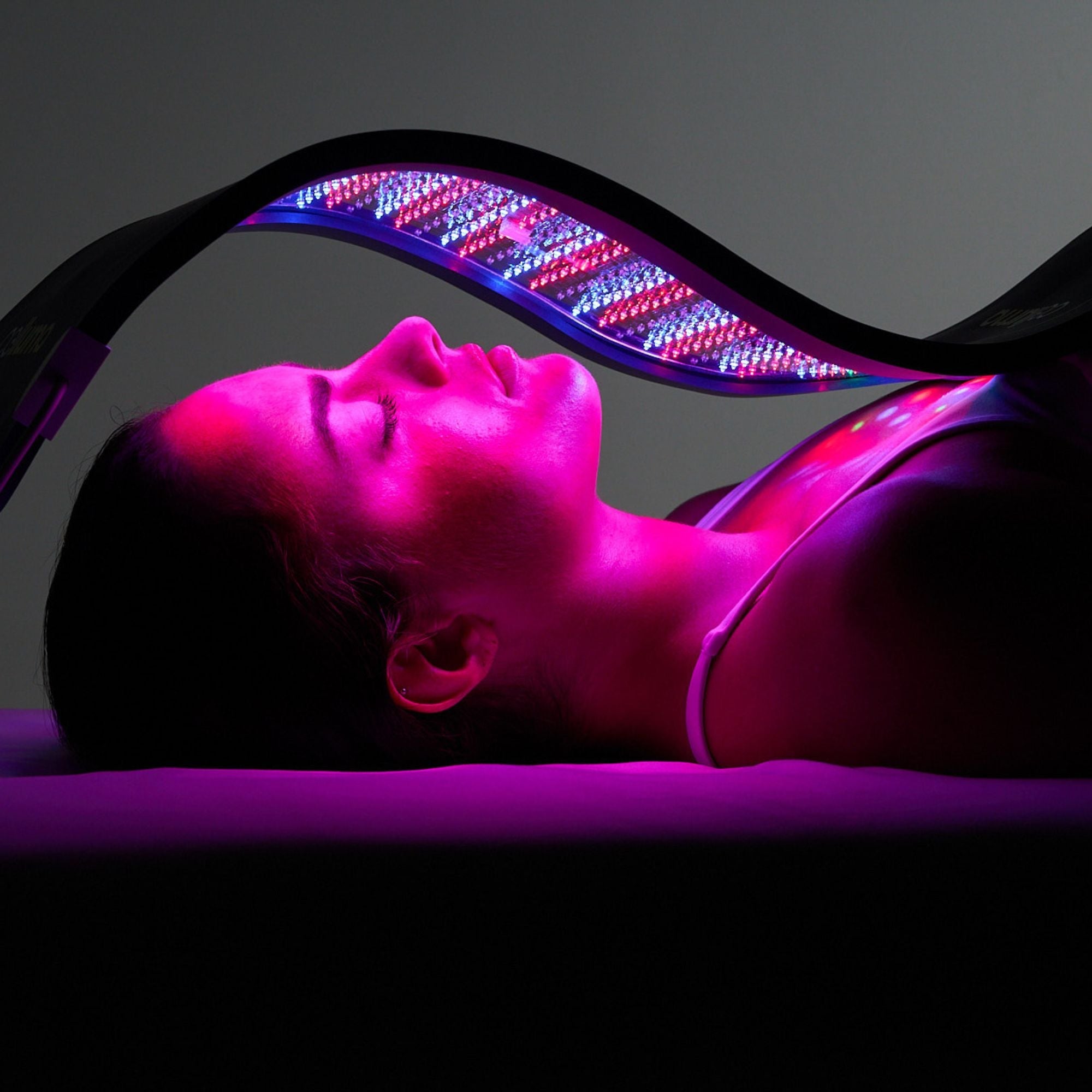 A person lies under a curved LED light therapy device emitting pink and blue light onto their face and neck.
