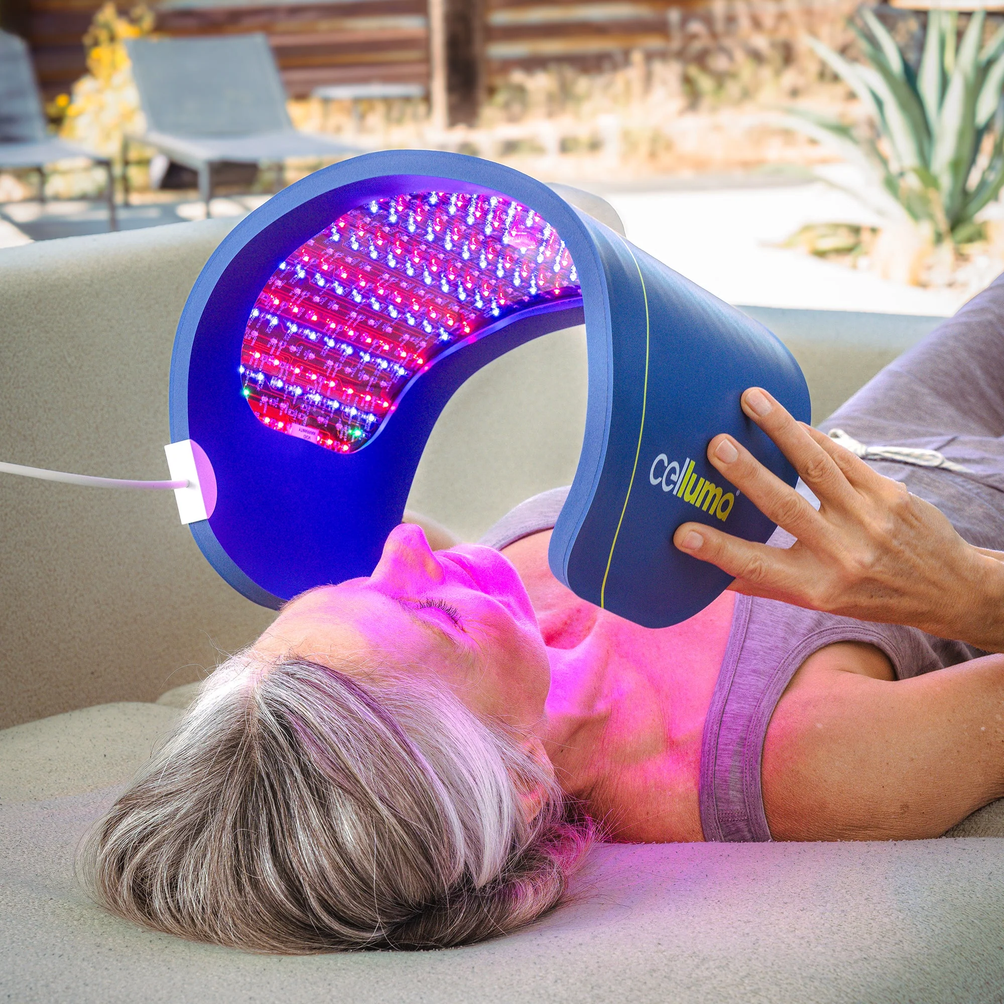 A person lies down with a curved LED light therapy device labeled "Celluma" positioned over their face, emitting red and blue light.