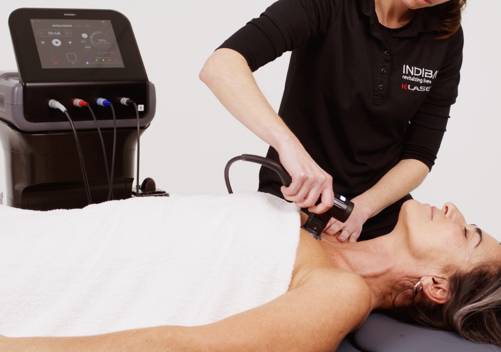 A woman lying on a treatment table is receiving a chest procedure with a handheld device operated by a technician next to a medical machine.