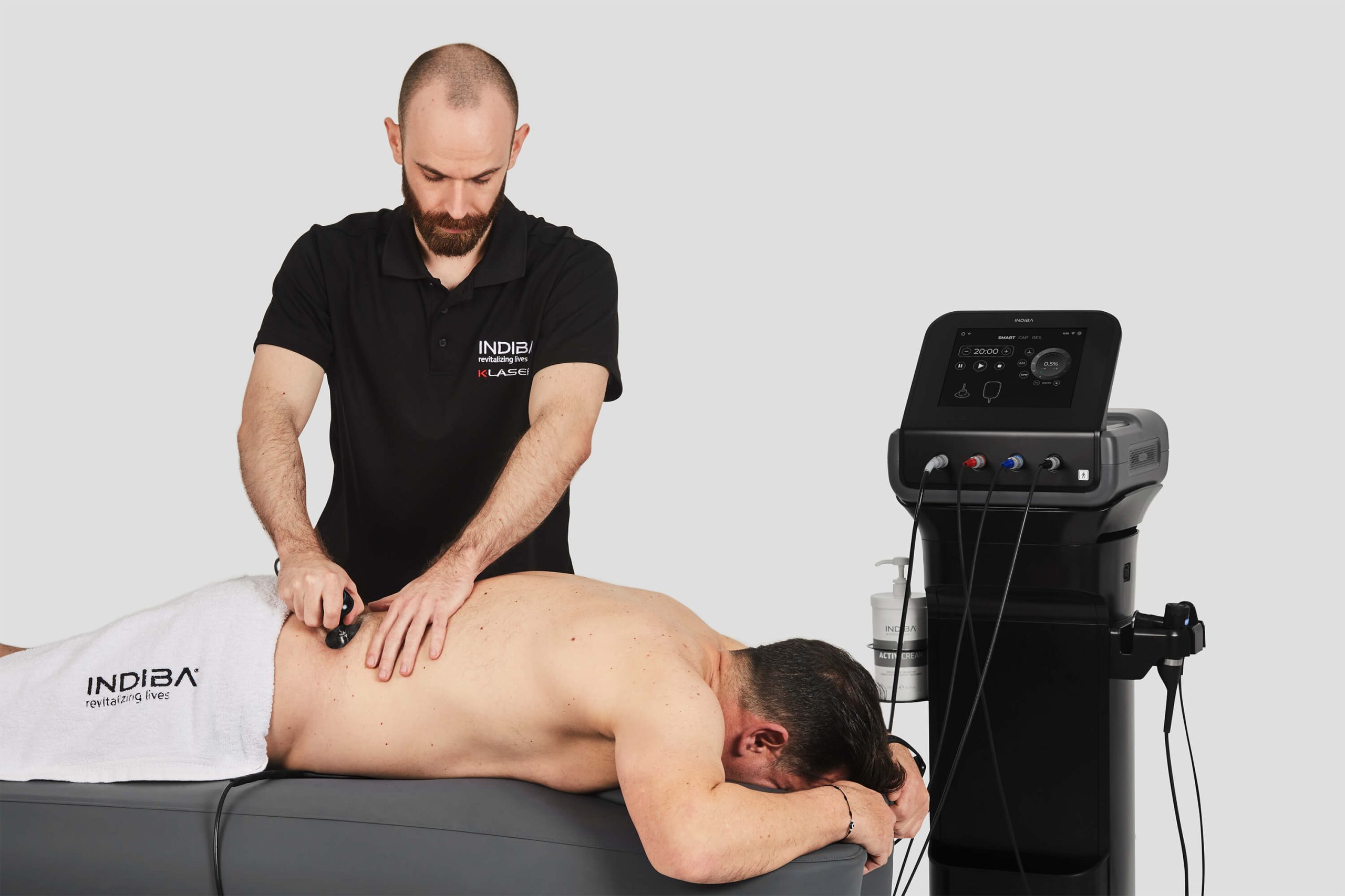 A therapist uses a handheld device on a man's bare back while he lies face down on a treatment table next to a medical machine.
